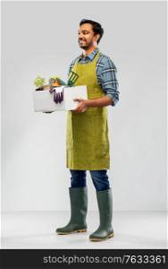 gardening, farming and people concept - happy smiling indian male gardener or farmer in apron and rubber boots with box of garden tools over grey background. indian gardener or farmer with box of garden tools