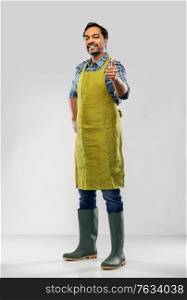 gardening, farming and people concept - happy smiling indian male gardener or farmer in apron and rubber boots showing thumbs up over grey background. indian male gardener or farmer showing thumbs up
