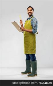 gardening, farming and people concept - happy smiling indian male gardener or farmer in apron and rubber boots with clipboard and pencil over grey background. happy indian gardener or farmer with clipboard