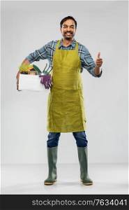 gardening, farming and people concept - happy smiling indian male gardener or farmer in apron and rubber boots with box of garden tools showing thumbs up over grey background. indian gardener or farmer with box of garden tools