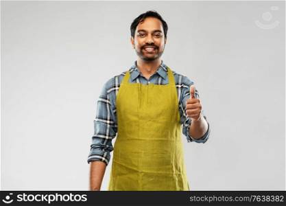 gardening, farming and people concept - happy smiling indian male gardener or farmer in apron showing thumbs up over grey background. indian male gardener or farmer showing thumbs up