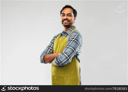 gardening, farming and people concept - happy smiling indian male gardener or farmer in apron with crossed arms over grey background. happy indian male gardener or farmer in apron