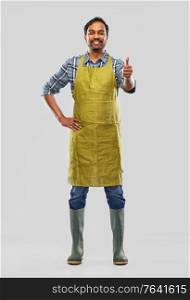 gardening, farming and people concept - happy smiling indian male gardener or farmer in apron and rubber boots showing thumbs up over grey background. indian male gardener or farmer showing thumbs up