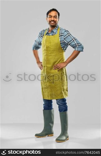 gardening, farming and people concept - happy smiling indian male gardener or farmer in apron and rubber boots over grey background. happy indian male gardener or farmer in apron