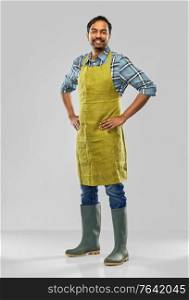gardening, farming and people concept - happy smiling indian male gardener or farmer in apron and rubber boots over grey background. happy indian male gardener or farmer in apron