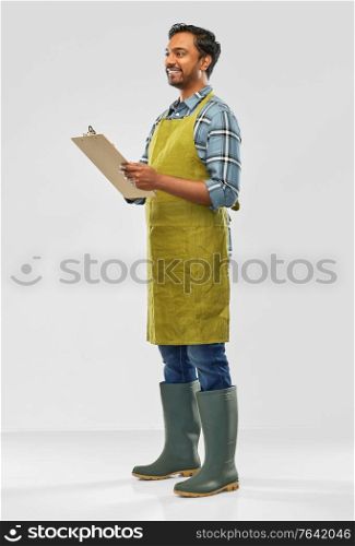 gardening, farming and people concept - happy smiling indian male gardener or farmer in apron and rubber boots with clipboard over grey background. happy indian gardener or farmer with clipboard