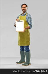 gardening, farming and people concept - happy smiling indian male gardener or farmer in apron and rubber boots with clipboard over grey background. happy indian gardener or farmer with clipboard