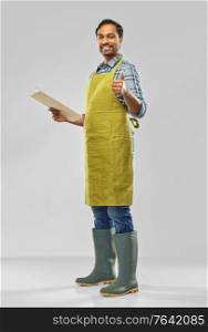 gardening, farming and people concept - happy smiling indian male gardener or farmer in apron and rubber boots with clipboard showing thumbs up over grey background. happy indian gardener or farmer with clipboard