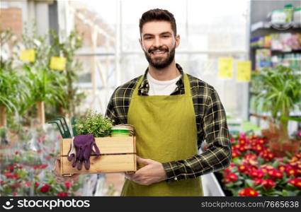 gardening, farming and people concept - happy smiling male gardener or seller in apron with box of garden tools over flower shop background. happy gardener or seller with box of garden tools