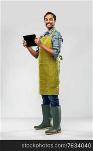 gardening, farming and technology concept - happy smiling indian male gardener or farmer in apron and rubber boots with tablet pc computer over grey background. indian male gardener or farmer with tablet pc