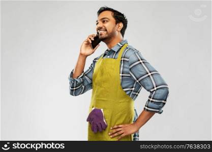 gardening, farming and technology concept - happy smiling indian male gardener or farmer in apron calling on smartphone over grey background. indian gardener or farmer calling on smartphone