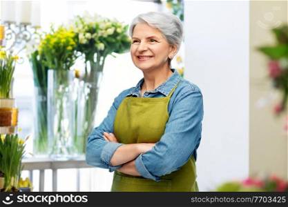 gardening, floristics and old people concept - portrait of smiling senior woman in green garden with apron crossed arms over flower shop background. portrait of smiling senior woman in garden apron