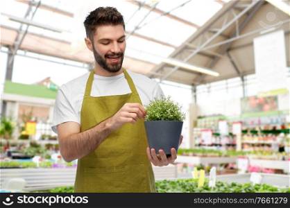 gardening, floristry and people concept - happy smiling male gardener in apron with flower in pot over store’s greenhouse background. smiling male gardener with flower in pot at shop