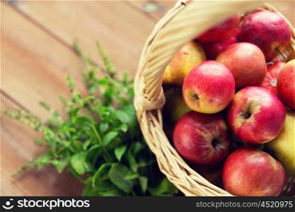 gardening, season, autumn and fruits concept - close up of wicker basket with ripe red apples and herbs on wooden table