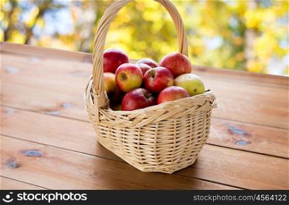 gardening, season, autumn and fruits concept - close up of wicker basket with ripe red apples on wooden table over natural background