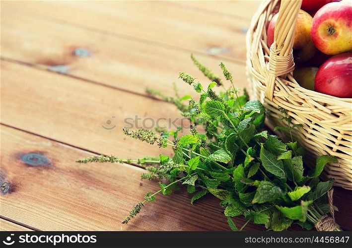 gardening, season, autumn, herbs and fruits concept - close up of wicker basket with ripe red apples and melissa bunch on wooden table