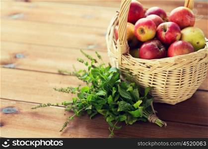 gardening, season, autumn, herbs and fruits concept - close up of wicker basket with ripe red apples and melissa bunch on wooden table