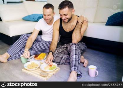Gay couple eating together sitting on their living room floor. Homosexual lifestyle concept.. Gay couple eating together sitting on their living room floor.