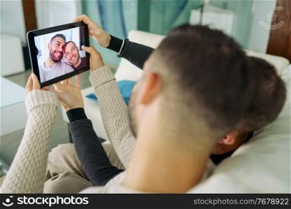 Gay couple making a selfie with a digital tablet sitting on the couch at home. Homosexual Lifestyle concept.. Gay couple making a selfie with a digital tablet sitting on the couch at home.