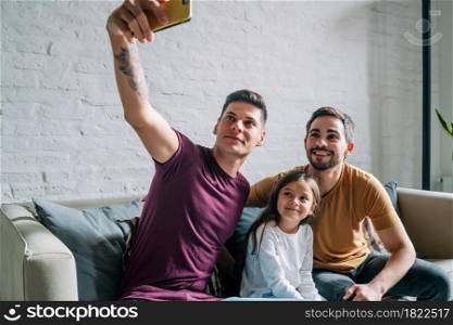 Gay couple taking a selfie with their daughter while sitting on a couch together at home. Family concept.