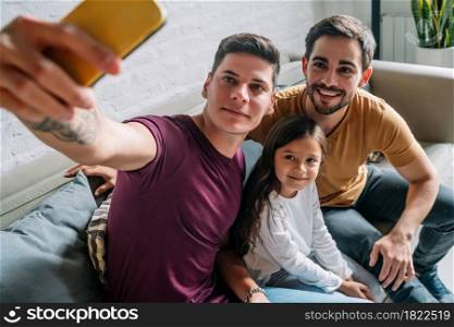Gay couple taking a selfie with their daughter while sitting on a couch together at home. Family concept.