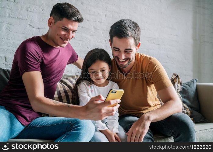 Gay couple using a mobile phone while sitting on a couch with their daughter at home. Family concept.