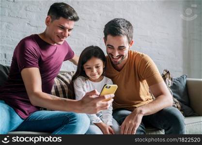 Gay couple using a mobile phone while sitting on a couch with their daughter at home. Family concept.