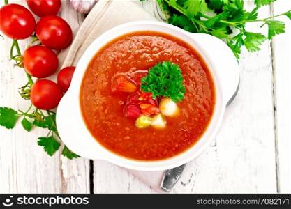 Gazpacho tomato soup in a white bowl with parsley and vegetables, spoon on a napkin against the background of the wooden planks on top