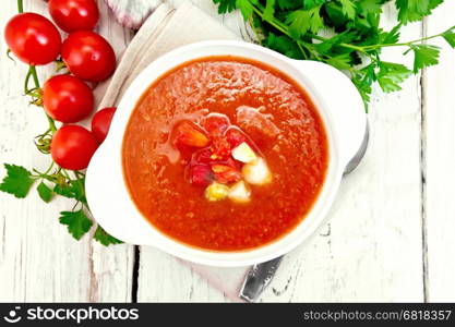 Gazpacho tomato soup in a white bowl with vegetables, spoon on a napkin, garlic and parsley on a background of wooden boards on top