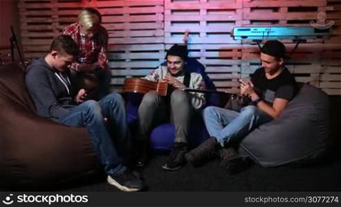 Generation Z musicians sitting on bean bag chairs on the stage using their mobile phones and chatting against musical instruments background. Music band relaxing after practice and training.
