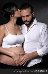 Gentle couple awaiting baby, beautiful pregnant woman kiss her husband over black background, holding hands on tummy in heart shape, happy anticipation of new life