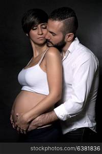 Gentle couple awaiting baby, beautiful pregnant woman with husband hugging with closed eyes over black background, happy anticipation of new life