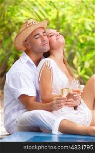 Gentle couple with closed eyes of pleasure sitting near the pool on tropical resort, drinking champagne on the beach, romantic summer vacation