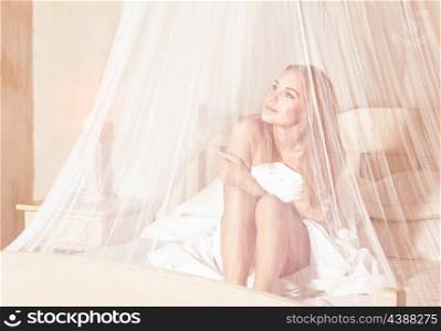Gentle girl enjoying wonderful day in luxury spa hotel, sitting on the bed under white transparent net and dreamy looking in side