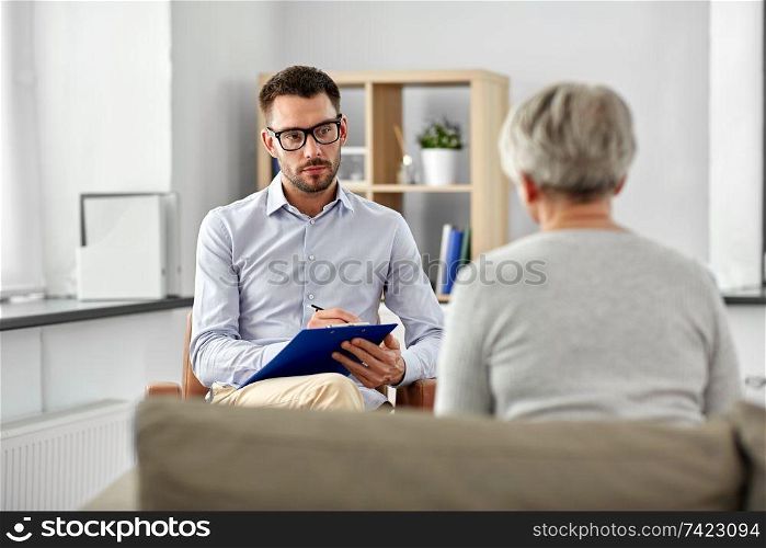 geriatric psychology, mental therapy and old age concept - psychologist with clipboard taking notes and listening to senior woman patient at psychotherapy session. psychologist listening to senior woman patient