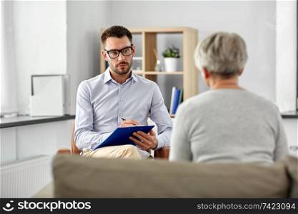 geriatric psychology, mental therapy and old age concept - psychologist with clipboard taking notes and listening to senior woman patient at psychotherapy session. psychologist listening to senior woman patient