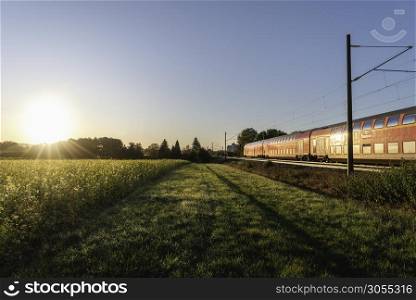 German red train moving on railway at sunrise, in spring scenery. Regional train and yellow flower field. Rapeseed and traveling train. Moving train.