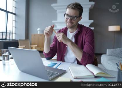 German young positive male entrepreneur in glasses having pleasant conversation with business partner by video call on laptop, smiling office worker discussing work results during online meeting. German young male entrepreneur in glasses having pleasant online conversation with colleagues
