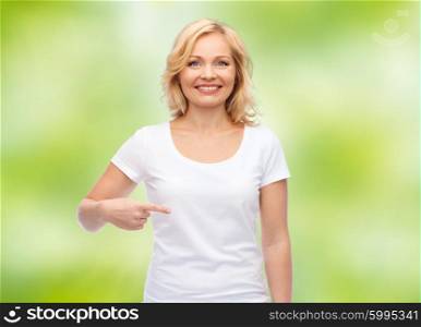 gesture, advertisement and people concept - smiling middle aged woman in blank white t-shirt pointing finger to herself over green natural background