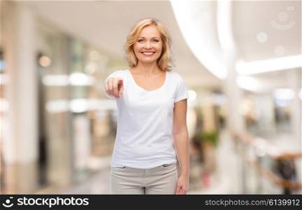 gesture, advertisement and people concept - smiling woman in blank white t-shirt pointing finger to you at shopping center or mall