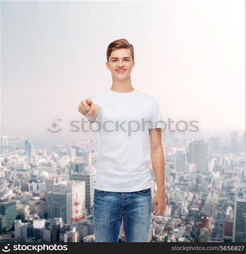 gesture, advertising and people concept - smiling young man in blank white t-shirt pointing at you over city background