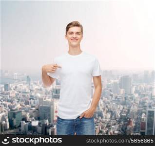 gesture, advertising and people concept - smiling young man in blank white t-shirt pointing finger on himself over city background