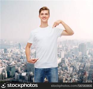 gesture, advertising and people concept - smiling young man in blank white t-shirt pointing fingers on himself over city background
