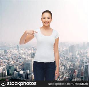 gesture, advertising and people concept - smiling young woman in blank white t-shirt pointing finger on herself over city background