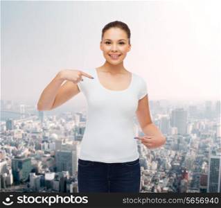 gesture, advertising and people concept - smiling young woman in blank white t-shirt pointing fingers on herself over city background