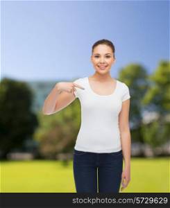 gesture, advertising, summer vacation and people concept - smiling young woman in blank white t-shirt pointing finger on herself over park background