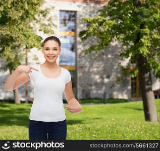 gesture, advertising, summer vacation, education and people concept - smiling young woman in blank white t-shirt pointing fingers on herself over campus background