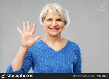 gesture and old people concept - portrait of smiling senior woman in blue sweater showing palm or five fingers over grey background. smiling senior woman showing palm or five fingers