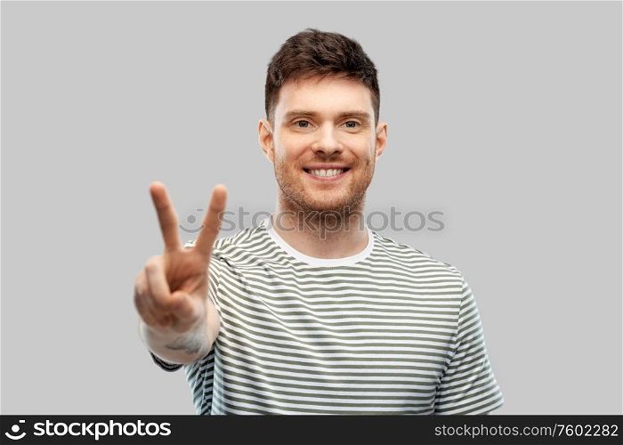 gesture and people concept - happy smiling young man in striped t-shirt showing peace hand sign over grey background. young man showing peace over grey background