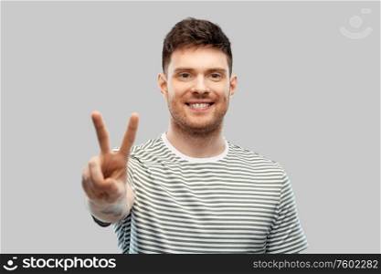 gesture and people concept - happy smiling young man in striped t-shirt showing peace hand sign over grey background. young man showing peace over grey background